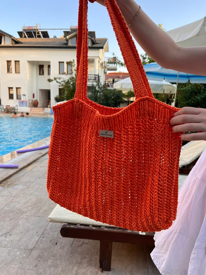 May include: A bright orange crochet tote bag featuring a vertical ribbed stitch pattern and two long shoulder straps. A small rectangular metal tag with the text "le handmade" is attached to the center front of the bag.