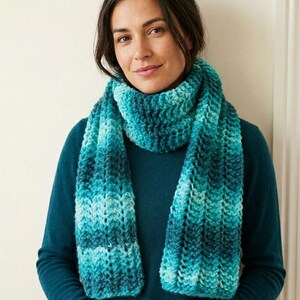 Op de afbeelding: Een grof gebreide sjaal met een gevarieerd kleurpatroon in tinten blauwgroen, turquoise en donkerblauw. De sjaal heeft een dikke, geribbelde textuur en is om de nek gewikkeld, met twee lange uiteinden die voor een donkerblauwe trui hangen.