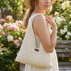 May include: A cream-coloured crocheted shoulder bag featuring a textured waffle stitch pattern, a single wide strap, and a small rectangular brown leather-like accent tag attached to the side of the bag near the top opening.