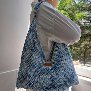 Op de afbeelding: Een grote, handgemaakte gehaakte tas in blauw- en grijstinten, met een bruin leren label. De tas heeft een lange riem en een driehoekige vorm. De tas is gemaakt van zacht garen.