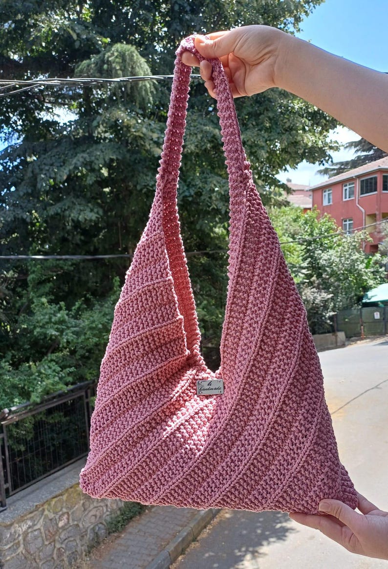 Pu&ograve; includere: Una borsa hobo all'uncinetto color rosa polvere con un motivo a coste diagonali e una tracolla integrata. Una piccola etichetta rettangolare in metallo color argento &egrave; fissata al centro sul davanti con il testo inciso "Handmade".