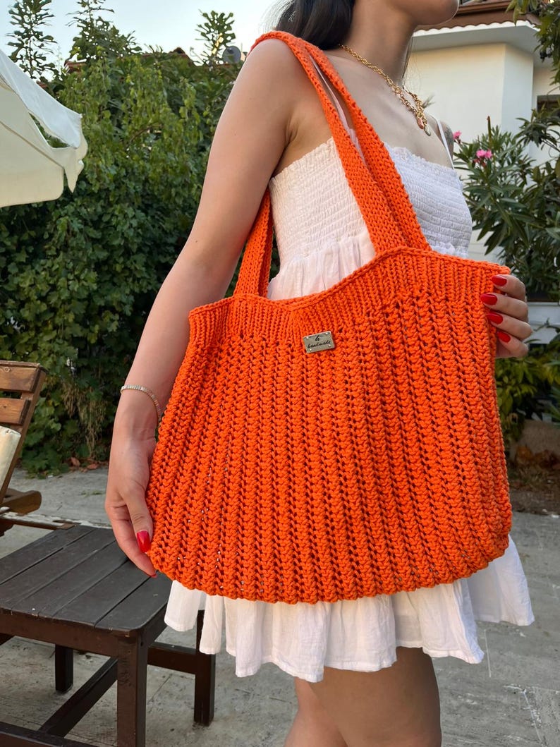 May include: A bright orange crochet tote bag featuring a vertical ribbed texture and two shoulder straps. A small rectangular metallic label with the text "Handmade" is attached to the center front of the bag, which is constructed from yarn.