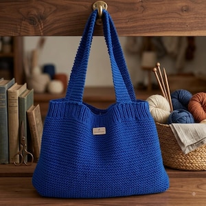 Op de afbeelding: Een levendige koningsblauwe gebreide tas met een getextureerd ribbelsteekpatroon, twee stevige schouderbanden en een klein rechthoekig beige label in het midden aan de voorzijde met de handgeschreven tekst "handmade" in zwarte inkt.