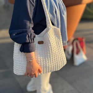 May include: A cream-coloured, hand-knitted tote bag with a rectangular shape and a textured pattern. The bag has a brown leather tag and sturdy straps. The bag is held by a person wearing a navy blue shirt and white trousers.