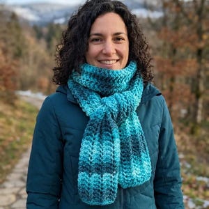 May include: A chunky, hand-crocheted scarf featuring a gradient of teal, turquoise, and dark blue yarn. The scarf has a ribbed, textured pattern and is wrapped around the neck, with two ends hanging down over a dark teal, zip-up hooded jacket.