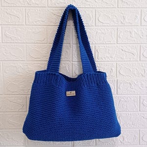May include: A striking blue knitted tote bag with strong handles, set against a white brick wall. The bag has a small rectangular label that reads "handmade". This handmade bag is ideal for carrying shopping or personal belongings.
