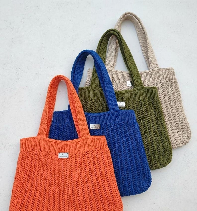 May include: A set of four crochet tote bags in orange, blue, olive green, and beige. Each bag features a vertical ribbed stitch pattern, two sturdy handles, and a small rectangular silver-toned metal tag attached to the front center.