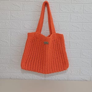 May include: A bright orange crochet tote bag featuring a vertical ribbed texture and two sturdy shoulder straps. A small rectangular label is attached to the center front of the bag, providing a subtle detail to the vibrant, hand-crafted design.