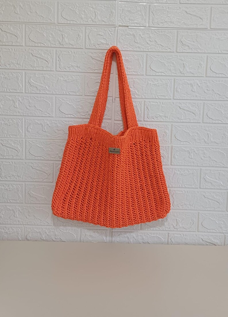 May include: A bright orange crochet tote bag featuring a vertical ribbed texture and two sturdy shoulder straps. A small rectangular label is attached to the center front of the bag, providing a subtle detail to the vibrant, hand-crafted design.