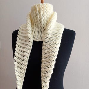 Può includere: Sciarpa color crema lavorata a mano, esposta su un manichino. La sciarpa presenta un design strutturato a trama aperta con un bordo smerlato. È drappeggiata attorno al collo del manichino, mostrando la sua forma triangolare e il suo aspetto accogliente.