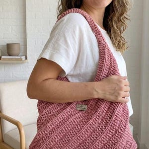 Pu&ograve; includere: Una borsa a spalla all'uncinetto color rosa polvere con un motivo a punti diagonali testurizzati e una piccola targhetta metallica rettangolare con la parola handmade incisa sul davanti. La borsa presenta un ampio manico integrato.