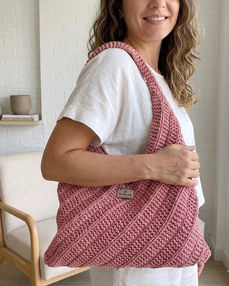 Pu&ograve; includere: Una borsa a spalla all'uncinetto color rosa polvere con un motivo a punti diagonali testurizzati e una piccola targhetta metallica rettangolare con la parola handmade incisa sul davanti. La borsa presenta un ampio manico integrato.
