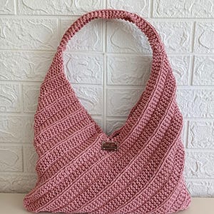 Peut inclure: Un sac fourre-tout en crochet rose poudré avec une seule bandoulière. Le sac présente un motif côtelé diagonal et une petite étiquette rectangulaire en bas. Le sac est posé contre un mur de briques blanches.