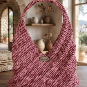 May include: A dusty rose crochet hobo bag featuring a diagonal ribbed texture and a single integrated handle. A small rectangular metal tag with the word handmade is centred on the front of the bag, which has a slouchy, unstructured silhouette.