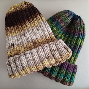 May include: Two knitted beanies with folded cuffs. One beanie features a variegated yarn pattern in shades of dark brown, tan, and cream. The second beanie is crafted from a multi-coloured yarn blend displaying vibrant hues of green, blue, and purple.