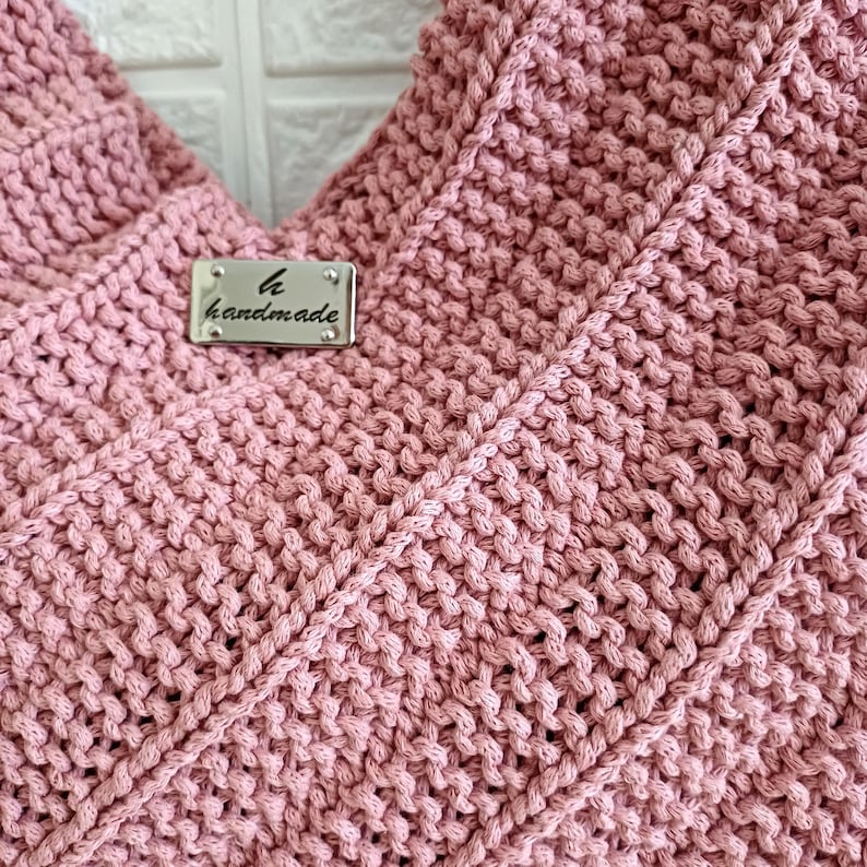 Pu&ograve; includere: Una borsa lavorata a maglia color rosa polvere con un motivo a legaccio testurizzato. Sulla parte anteriore &egrave; fissata un'etichetta rettangolare in metallo argentato, incisa con la lettera h e la parola handmade in carattere corsivo.