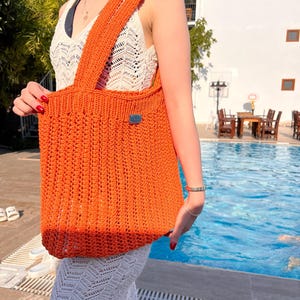 May include: A bright orange crochet tote bag featuring a vertical ribbed texture and a small rectangular metallic brand tag on the front. The bag has two wide shoulder straps and is constructed from a thick, durable yarn in a solid vibrant hue.