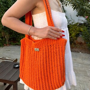 May include: A bright orange crochet tote bag featuring a vertical ribbed stitch pattern and two shoulder straps. A small rectangular wooden tag with the text "handmade" is attached to the upper front center of the bag, which is made of yarn.