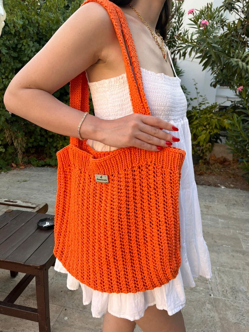 May include: A bright orange crochet tote bag featuring a vertical ribbed stitch pattern and two shoulder straps. A small rectangular wooden tag with the text "handmade" is attached to the upper front center of the bag, which is made of yarn.