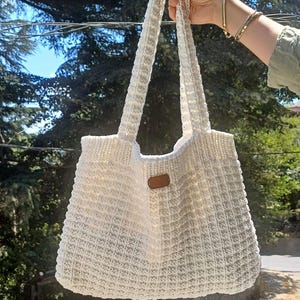 May include: A white crocheted tote bag featuring a textured waffle-like stitch pattern and two long shoulder straps. A small, rectangular brown leather-like tag is centred on the front of the bag, providing a subtle contrast to the white yarn.