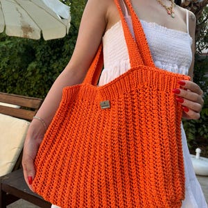 Peut inclure: Sac fourre-tout en crochet orange vif avec de larges bretelles. Le sac est de forme rectangulaire et comporte une petite étiquette rectangulaire. Fabriqué à partir de fil épais, il présente un aspect texturé. Le sac est tenu par une personne portant une robe blanche.
