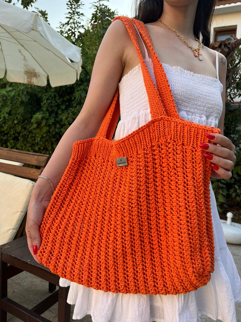 May include: A bright orange crochet tote bag featuring a textured vertical ribbed pattern and two shoulder straps. A small rectangular metallic brand tag is attached to the front center of the bag, providing a subtle contrast to the vibrant yarn.
