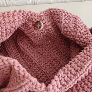 Pu&ograve; includere: Una borsa lavorata a maglia color rosa polvere con un motivo a legaccio testurizzato, una chiusura magnetica circolare color argento sull'apertura superiore e una placca metallica rettangolare color argento fissata sulla superficie esterna anteriore.