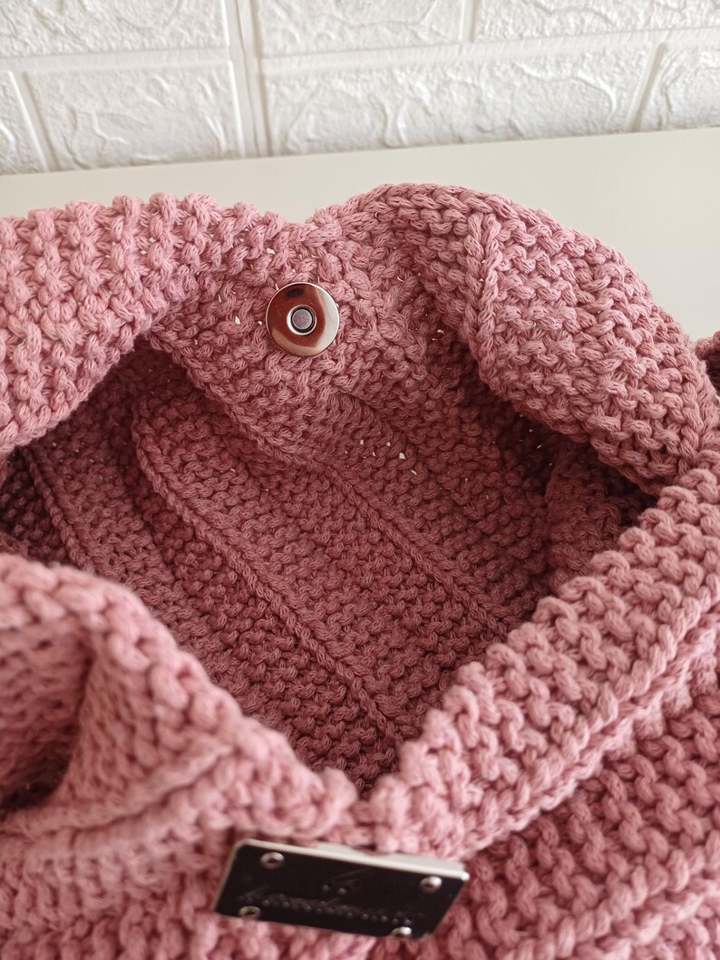 Pu&ograve; includere: Una borsa lavorata a maglia color rosa polvere con un motivo a legaccio testurizzato, una chiusura magnetica circolare color argento sull'apertura superiore e una placca metallica rettangolare color argento fissata sulla superficie esterna anteriore.