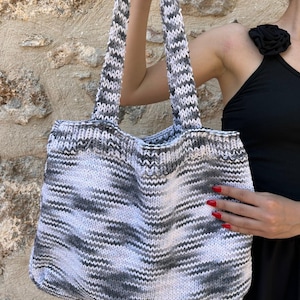 May include: A knitted tote bag with a zig-zag pattern in shades of gray and white. The bag has two thick, matching handles. The bag is held up by a person.