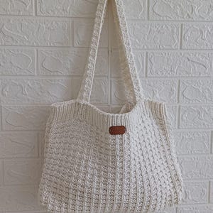 May include: A cream-coloured, knitted tote bag with long handles. The bag has a textured stitch pattern and a small, brown leather tag. It hangs against a white brick wall.