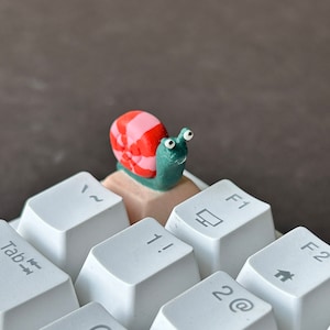 May include: A small, green and pink snail figurine sits on a white computer keyboard. The snail has googly eyes and a red and white striped shell. The keyboard has keys labeled "Tab", "~", "F1", "F2", "1!", and "2@."