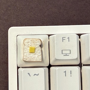 May include: A custom keyboard keycap featuring a slice of toast with a square of yellow butter. The keycap is a replacement for the Esc key and is surrounded by other white keys with black lettering.