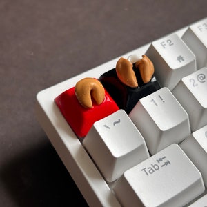 Puede incluir: Dos teclas de teclado personalizadas con diseños de galletas de la fortuna. Las teclas son rojas y negras con una galleta de la fortuna dorada en la parte superior de cada una. Las teclas están en un teclado blanco.