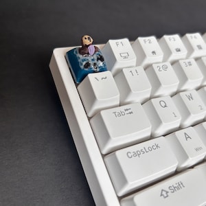 May include: A white mechanical keyboard with a custom keycap featuring a small figurine of a person wearing a suit and tie. The keycap is blue and has a textured surface.
