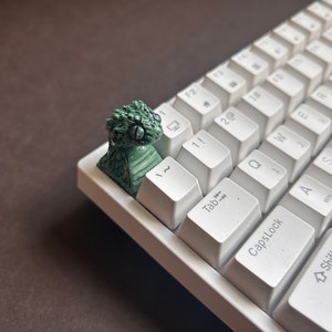 Può includere: Una tastiera bianca con un copritasto di drago verde sulla chiave di escape. La tastiera ha uno sfondo bianco e i tasti sono bianchi con lettere nere.