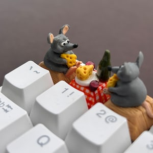 Può includere: Due figurine di topi grigi sono seduti su una tastiera bianca, mangiando formaggio e bevendo vino. I topi sono seduti sopra i tasti etichettati "1!" e "2@" con un piccolo tovagliolo a scacchi rosso e bianco tra loro.
