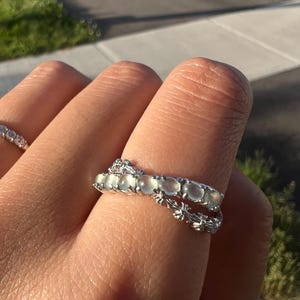 Könnte beinhalten: Ein silberner Ring mit einer Reihe ovaler, hellgrüner Edelsteine. Der Ring wird an einem Finger getragen, darunter ein zweiter Ring mit floralem Design. Der Hintergrund zeigt eine sonnige Szene im Freien.