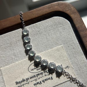 May include: A silver chain bracelet with a row of round, light green jade stones set in silver. Each stone is surrounded by a halo of smaller, clear stones. The bracelet is lying on a white fabric surface with a label that reads "French Paris, the dead tree of spring".