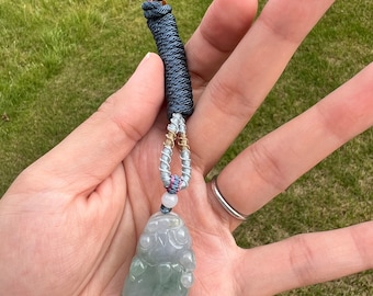 Puede incluir: Un colgante de jade verde en forma de criatura mítica, colgado de un cordón plateado y azul con una cuenta negra en la parte superior.