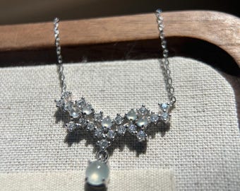 May include: A delicate silver necklace featuring a floral design with small, clear gemstones and a larger, oval-shaped pendant. The necklace is displayed on a neutral-toned surface, showcasing its intricate details and elegant style.