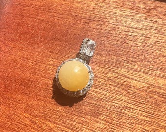 Puede incluir: Un colgante de piedra amarilla con una piedra redonda engastada en bisel rodeada de piedras transparentes más pequeñas. El colgante está unido a una cadena de plata con un engaste de piedra transparente.