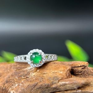 May include: A silver ring with a green gemstone surrounded by a halo of smaller white gemstones. The ring is resting on a piece of wood.