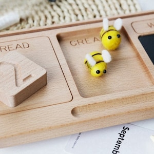 May include: Wooden tray with four sections labeled "Read", "Create", "Write", and a blank section. The tray includes a wooden number two, two felt bee figurines, and a small chalkboard.