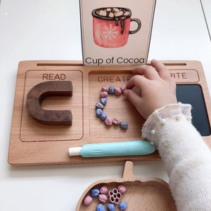 May include: A wooden learning tray with three sections labeled "Read", "Create", and "Write". The "Create" section has a child's hand placing small pink and blue beads in the shape of the letter "C". A card with the letter "Cc" and a picture of a red mug with hot chocolate and marshmallows is in the top section of the tray.