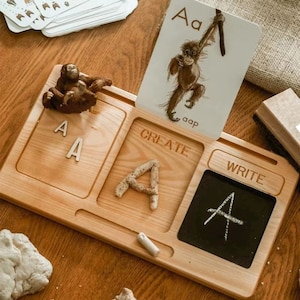 May include: Wooden learning tray with three sections labeled "Create", "Write", and "A". The tray has a small chalkboard, a space for play dough, and a space for letter tiles. There is a small toy orangutan and a flashcard with the letter "A" and a picture of an orangutan.