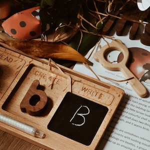 May include: Wooden learning toy with three sections labeled "Read", "Create", and "Write". The "Create" section has a wooden letter "B" and the "Write" section has a small chalkboard with a white chalk letter "B" written on it. A white chalk stick is lying next to the chalkboard.