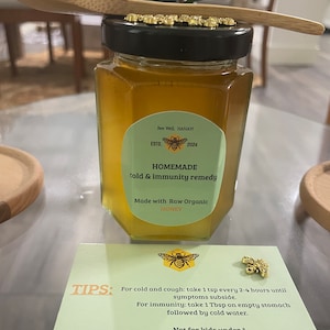 May include: A glass jar of honey with a green label that reads "Bee Well HANAYI ESTD. 2024 HOMEMADE cold & immunity remedy Made with Raw Organic HONEY". The jar is sitting on a wooden surface with a wooden spoon on top. A small card with the text "TIPS: For cold and cough: take 1 tsp every 2-4 hours until symptoms subside. For immunity: take 1 Tbsp on empty stomach followed by cold water. Not for kids under 1." is in the foreground.