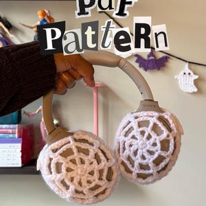 Può includere: Cuffie Sony con copriauricolari all'uncinetto rosa chiaro. Il testo "Pdf Pattern" è visualizzato in alto e "By: Stitched by Soleil" in basso. Decorazioni di Halloween all'uncinetto sullo sfondo.