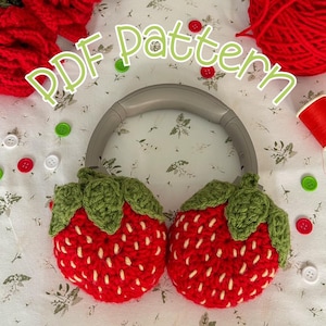 Puede incluir: Calentadores de orejas de fresa de ganchillo en rojo y verde, con una diadema gris. Los calentadores de orejas tienen forma de fresas con hojas verdes. El texto "PDF pattern" está escrito en letras verdes sobre un fondo blanco.