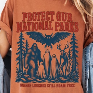 May include: A rust-colored t-shirt featuring a dark blue graphic of a forest scene with Bigfoot, ghosts, a deer-like creature, and an owl. The text reads "PROTECT OUR NATIONAL PARKS" and "WHERE LEGENDS STILL ROAM FREE" in a bold, red, distressed font.